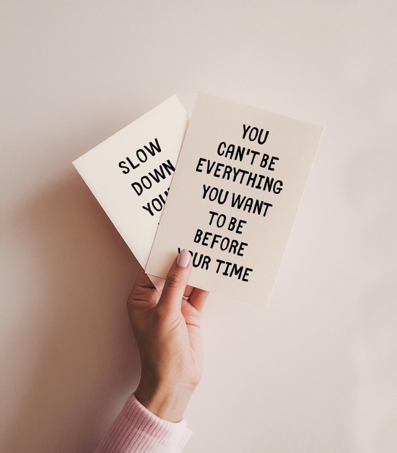 May include: Two cream-colored cards with black text are held in a hand. One card reads "SLOW DOWN YOU" and the other reads "YOU CAN'T BE EVERYTHING YOU WANT TO BE BEFORE YOUR TIME."