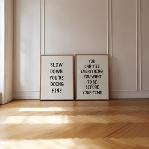 May include: Two framed art prints with motivational quotes. The left print reads "SLOW DOWN YOU'RE DOING FINE" and the right print says "YOU CAN'T BE EVERYTHING YOU WANT TO BE BEFORE YOUR TIME". The frames are brown, and the background is white.