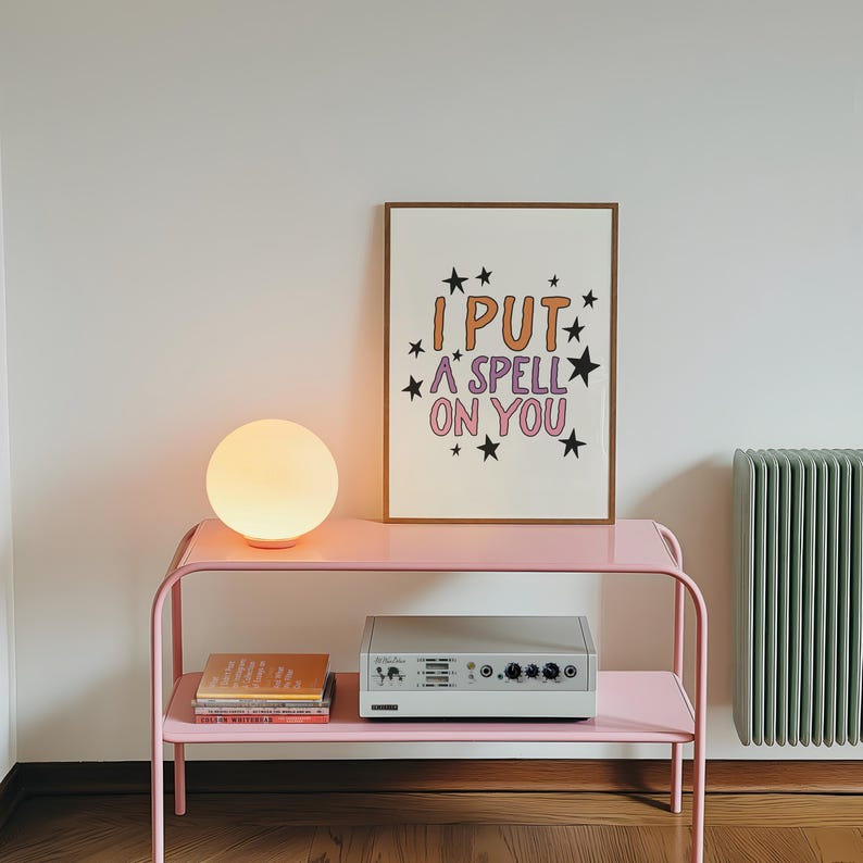 May include: A pink metal side table with a round, glowing lamp and a framed print that reads "I PUT A SPELL ON YOU." A silver audio device and books are on the lower shelf. The print has a brown frame.