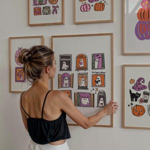 May include: A collection of framed Halloween-themed art prints. The prints feature illustrations of ghosts in windows, pumpkins, witch hats, and other spooky elements. The color palette includes orange, purple, black, and white, with wooden frames.