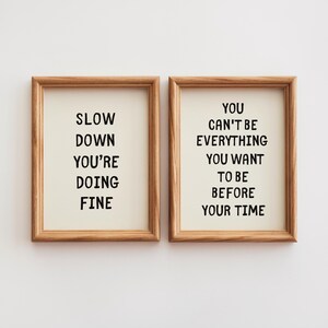 May include: Two framed art prints with inspirational quotes. One reads "SLOW DOWN YOU'RE DOING FINE" and the other says "YOU CAN'T BE EVERYTHING YOU WANT TO BE BEFORE YOUR TIME". The frames are made of light brown wood.