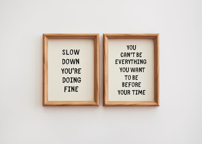 May include: Two framed art prints with inspirational quotes. One reads "SLOW DOWN YOU'RE DOING FINE" and the other says "YOU CAN'T BE EVERYTHING YOU WANT TO BE BEFORE YOUR TIME". The frames are made of light brown wood.