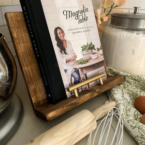May include: A brown wooden cookbook stand with a gold metal handle. A cookbook titled "Magnolia Table" by Joanna Gaines is displayed on the stand. A rolling pin and a whisk are on the counter.