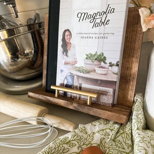 May include: A cookbook titled "Magnolia Table" by Joanna Gaines, featuring a collection of recipes for gathering. The book is displayed on a wooden stand with a gold metal handle.
