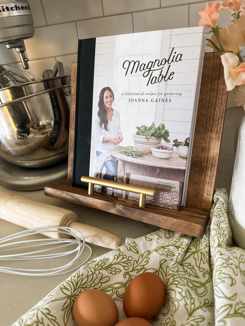May include: A cookbook titled "Magnolia Table" by Joanna Gaines, featuring a collection of recipes for gathering. The book is displayed on a wooden stand with a gold metal handle.
