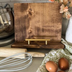 May include: A wooden recipe stand with a brown stain finish and a gold metal handle. The stand is sitting on a countertop with a rolling pin, whisk, and eggs on a patterned tea towel.