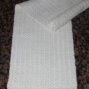 Peut inclure: Un chemin de table blanc tissé avec une surface texturée et des franges aux extrémités. Le chemin de table est plié en deux et posé sur une surface brune.