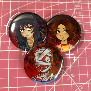 May include: Three round buttons with cartoon illustrations. One button features a mummy with bandages and a blue eye. Another depicts a person with dark hair and a striped shirt. The third shows a person with brown hair and a pink shirt.
