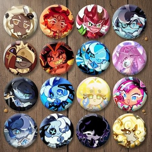 Puede incluir: Una colección de dieciséis botones redondos con diseños de personajes de dibujos animados coloridos. Los botones muestran una variedad de personajes con diferentes expresiones y combinaciones de colores, sobre un fondo de madera. Cada botón mide aproximadamente 3,8 cm de diámetro.