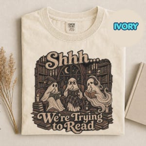 Może przedstawiać: Koszulka w kolorze kości słoniowej z grafiką przedstawiającą trzy duchy siedzące w bibliotece i czytające książki. Napis głosi "Shhh... We're Trying to Read". Ilustracja zawiera regały z książkami, świece i półksiężyc widoczny w oknie.