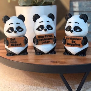 May include: Three panda figurines with black and white fur, each holding a brown sign. The signs read: "Eat. Nap. Repeat.", "Nap First. Decisions Later", and "I'd Rather Be Napping". The figurines are on a wooden tray.