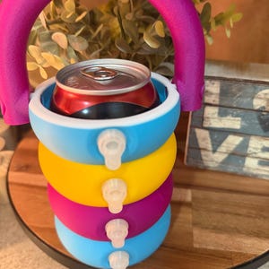 May include: A colorful, stackable can cooler with a pink handle. The cooler is made of blue, yellow, and magenta rings, each with a small white spout. A red can sits inside the top ring. The cooler is designed to keep beverages cold.