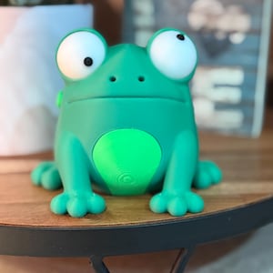 May include: A whimsical, green frog figurine with large, white-rimmed eyes and a round belly. The frog has a friendly expression and is sitting on a wooden surface. The figurine is a fun decorative item.