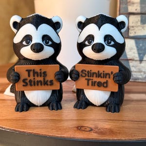 May include: Two raccoon figurines, black and white with orange signs. One sign reads "This Stinks" and the other says "Stinkin' Tired". The figurines are on a wooden surface.