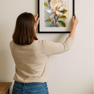 May include: A framed floral art print featuring a white magnolia blossom with yellow and green accents. The artwork is set against a soft, light blue background, framed in a black border. The print is being held up against a white wall.