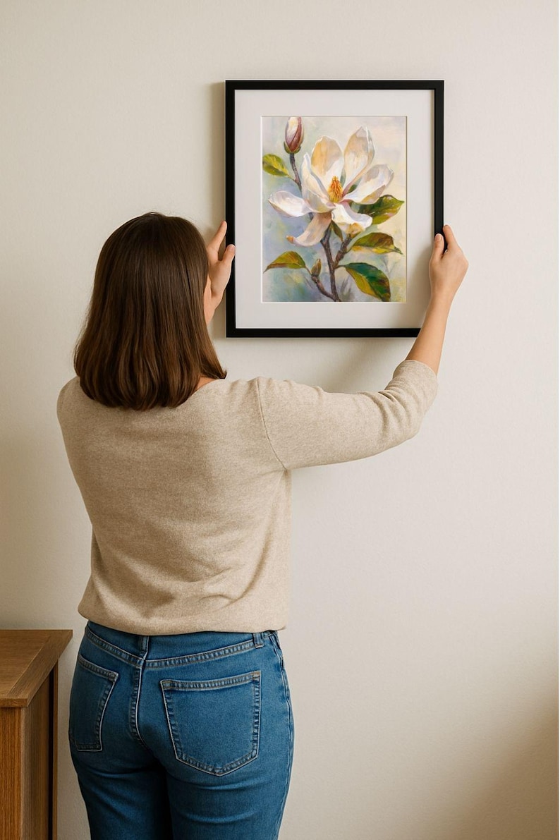 May include: A framed floral art print featuring a white magnolia blossom with yellow and green accents. The artwork is set against a soft, light blue background, framed in a black border. The print is being held up against a white wall.