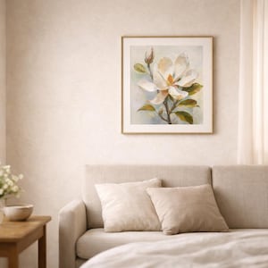 May include: A framed floral print featuring a large white magnolia blossom with yellow center and green leaves. The artwork is in a gold frame and hangs above a beige sofa with two matching pillows. The room has a neutral color palette.