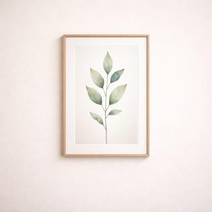 Puede incluir: Una impresión en acuarela enmarcada de una ramita botánica con hojas verdes. La obra de arte está sobre un fondo blanco y enmarcada en un marco de madera marrón claro. La pared detrás del marco es blanca.