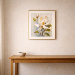 May include: A framed painting of a magnolia flower in full bloom, with a bud and green leaves. The artwork features soft colors of white, yellow, and green against a light blue background. The painting is displayed on a wall above a wooden table.