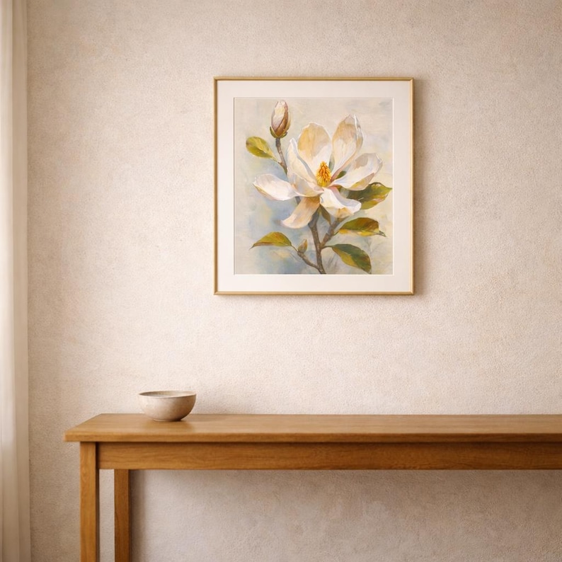 May include: A framed painting of a magnolia flower in full bloom, with a bud and green leaves. The artwork features soft colors of white, yellow, and green against a light blue background. The painting is displayed on a wall above a wooden table.