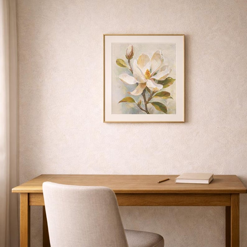 May include: A framed watercolor painting of a white magnolia flower with green leaves and a bud. The artwork is in a gold frame and hangs on a textured wall above a wooden desk. A chair, a book, and a pencil are also visible.