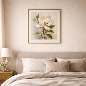May include: A framed floral print featuring a large white magnolia blossom with yellow center and green leaves. The artwork is in a gold frame and hangs above a bed with neutral-toned bedding.