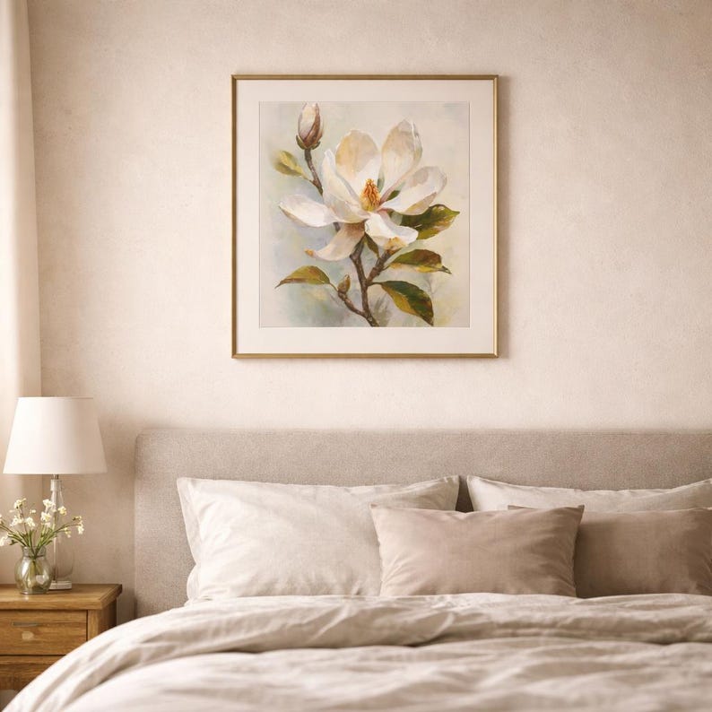 May include: A framed floral print featuring a large white magnolia blossom with yellow center and green leaves. The artwork is in a gold frame and hangs above a bed with neutral-toned bedding.