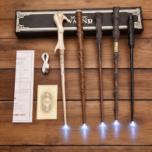 Pode incluir: Uma coleção de cinco varinhas mágicas detalhadas em tons de castanho e preto, cada uma com uma ponta branca brilhante. As varinhas são exibidas com uma caixa que diz "MAGIC WAND". Um cabo de carregamento e um pequeno cartão também estão presentes.