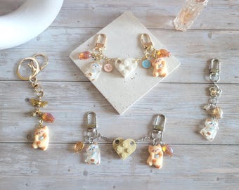Puede incluir: Una colección de llaveros con dijes decorativos. Los llaveros presentan pequeñas y detalladas figuras de osos y gatos, junto con detalles en forma de corazón y florales. La paleta de colores incluye dorado, crema y tonos pastel.
