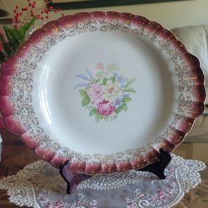May include: A large, white porcelain plate with a burgundy rim and gold accents. The plate features a floral design with pink, white, and blue flowers.
