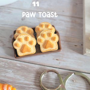 Puede incluir: Cuatro tostadas con forma de pata sobre una bandeja marr&oacute;n. La tostada es de color marr&oacute;n dorado con un dise&ntilde;o de huella de pata. El texto "11 Paw Toast" est&aacute; encima de la tostada. Un llavero plateado est&aacute; en primer plano.