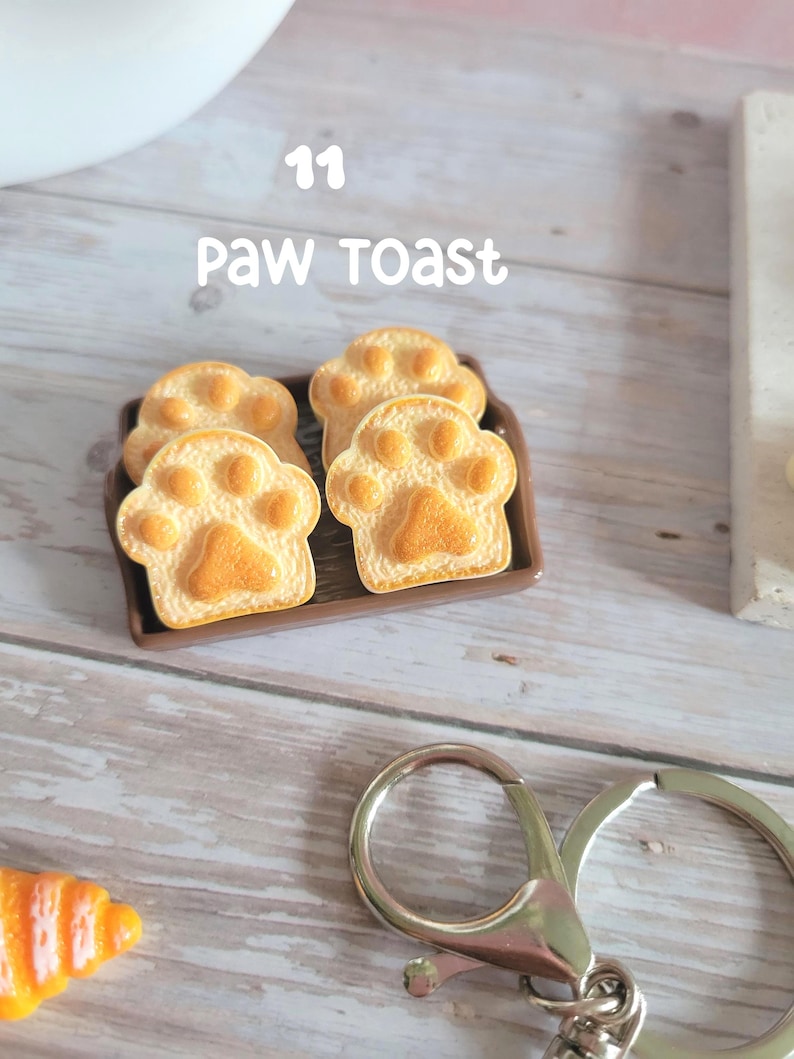 Puede incluir: Cuatro tostadas con forma de pata sobre una bandeja marr&oacute;n. La tostada es de color marr&oacute;n dorado con un dise&ntilde;o de huella de pata. El texto "11 Paw Toast" est&aacute; encima de la tostada. Un llavero plateado est&aacute; en primer plano.
