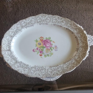 Puede incluir: Un plato ovalado blanco para servir con un diseño floral dorado y plateado. El plato tiene un borde festoneado y un patrón floral en el centro.