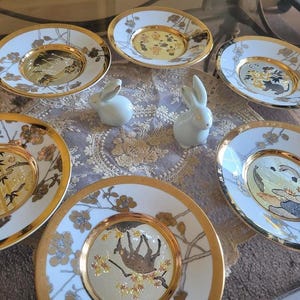 Op de afbeelding: Een verzameling van zes witte en gouden decoratieve borden met ingewikkelde Aziatisch geïnspireerde ontwerpen. Twee kleine witte keramische konijnenbeeldjes zijn ook aanwezig. De borden hebben gouden randen en gedetailleerde kunstwerken in het midden.