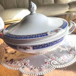 Pode incluir: Uma sopeira de porcelana branca com tampa, com uma borda decorativa azul e dourada com detalhes florais. A sopeira tem duas alças e um adorno decorativo na tampa. Está sobre uma toalha de renda branca.