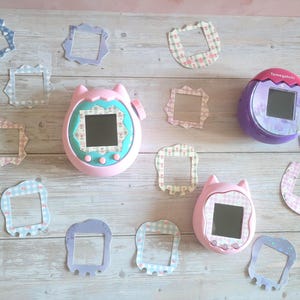 May include: A collection of Tamagotchi virtual pet devices in pink and purple, with decorative paper frames in pastel colors. The devices have screens and buttons, set against a light wood grain background.