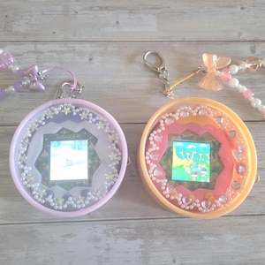 Peut inclure: Deux animaux de compagnie numériques Tamagotchi ronds avec des étuis décoratifs. L'appareil de gauche est lavande avec une sangle perlée violette et des étoiles. L'appareil de droite est pêche avec une sangle perlée rose et blanche et un nœud.