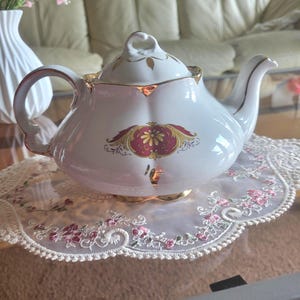 May include: A white porcelain teapot with gold detailing and a red and yellow floral pattern. The teapot rests on a white lace doily with pink floral embroidery. A white vase with pink flowers is in the background.