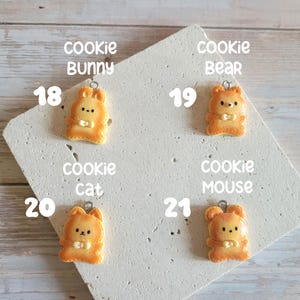 Puede incluir: Cuatro colgantes con forma de galleta de color naranja con forma de conejo, oso, gato y rat&oacute;n. Cada colgante tiene un peque&ntilde;o lazo plateado en la parte superior. Los colgantes est&aacute;n sobre una superficie blanca en &aacute;ngulo con las palabras "cookie bunny", "cookie bear", "cookie cat" y "cookie mouse".