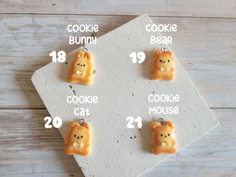 Puede incluir: Cuatro colgantes con forma de galleta de color naranja con forma de conejo, oso, gato y rat&oacute;n. Cada colgante tiene un peque&ntilde;o lazo plateado en la parte superior. Los colgantes est&aacute;n sobre una superficie blanca en &aacute;ngulo con las palabras "cookie bunny", "cookie bear", "cookie cat" y "cookie mouse".