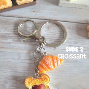 Puede incluir: Un llavero plateado con dos colgantes tem&aacute;ticos de comida. Un colgante es una rebanada de pan tostado con un centro en forma de coraz&oacute;n, y el otro es un cruas&aacute;n naranja. El texto "SIDE 2 CROISSANT" es visible.