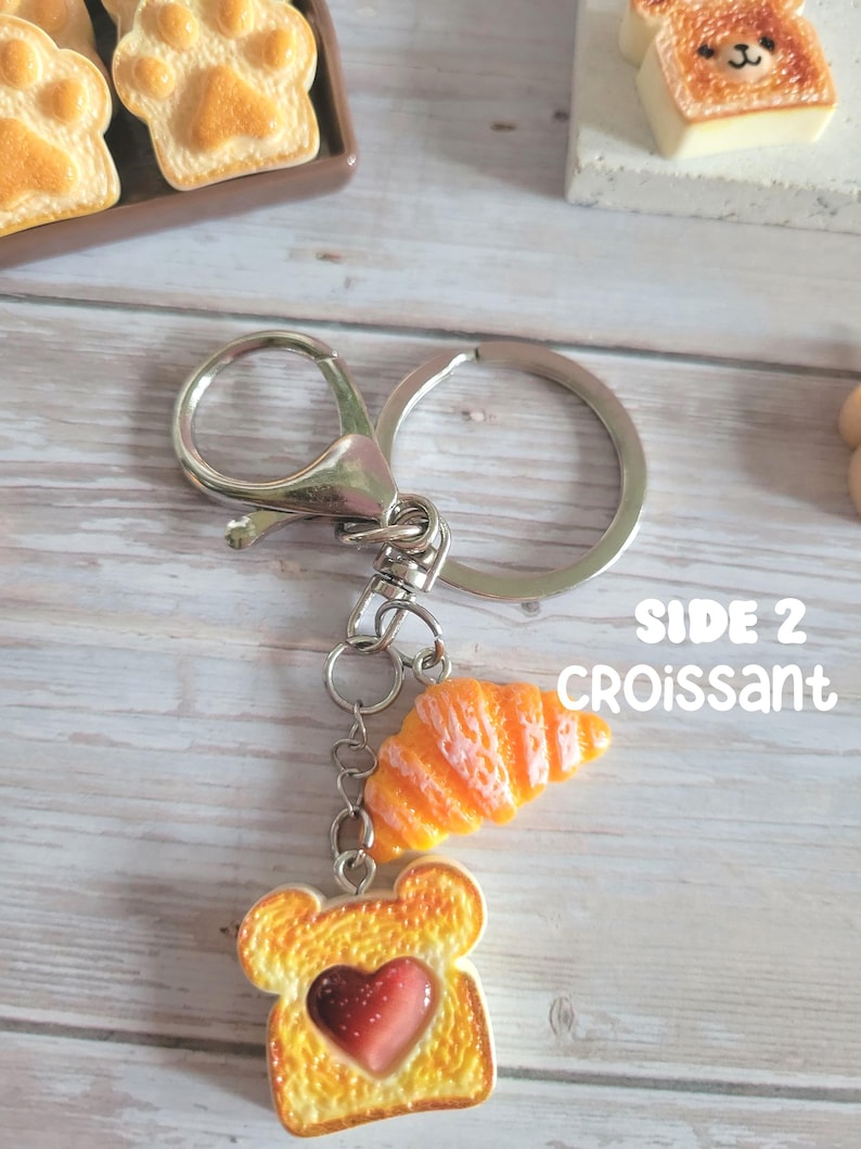 Puede incluir: Un llavero plateado con dos colgantes tem&aacute;ticos de comida. Un colgante es una rebanada de pan tostado con un centro en forma de coraz&oacute;n, y el otro es un cruas&aacute;n naranja. El texto "SIDE 2 CROISSANT" es visible.