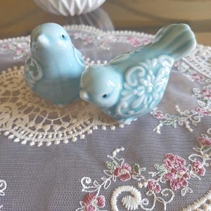 May include: Two pale blue ceramic bird ornaments with floral detailing. They sit on a lace doily with pink and green floral embroidery. A white, ribbed vase is in the background.