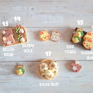 Puede incluir: Una colecci&oacute;n de colgantes en miniatura con tem&aacute;tica de comida, que incluyen un oso ruborizado, yema de conejo, tostada de huevo, conejo de fresa y panecillo de gatito. Los colgantes est&aacute;n hechos de arcilla polim&eacute;rica y est&aacute;n dispuestos sobre una superficie de madera clara.