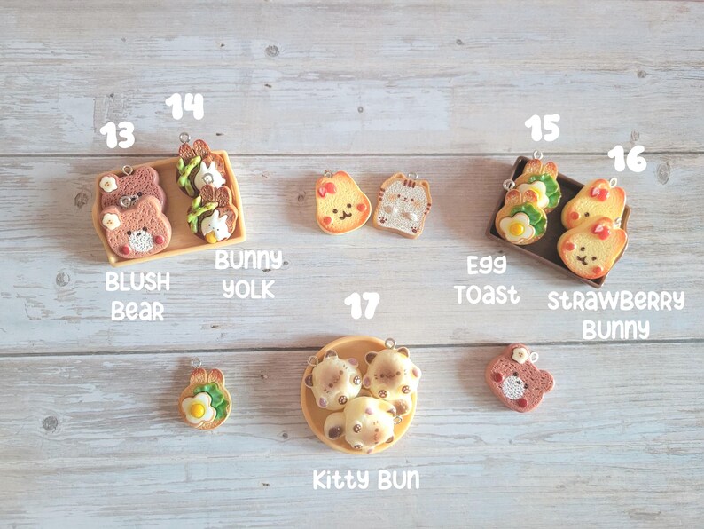 Puede incluir: Una colecci&oacute;n de colgantes en miniatura con tem&aacute;tica de comida, que incluyen un oso ruborizado, yema de conejo, tostada de huevo, conejo de fresa y panecillo de gatito. Los colgantes est&aacute;n hechos de arcilla polim&eacute;rica y est&aacute;n dispuestos sobre una superficie de madera clara.