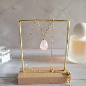 May include: A gold-colored necklace with a pear-shaped, iridescent pendant is displayed on a gold stand with a wooden base. The necklace chain is delicate, and the pendant has a soft, glowing appearance. The background is blurred, with a perfume bottle visible.