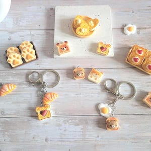 Puede incluir: Una variedad de colgantes y llaveros en miniatura con tem&aacute;tica de comida. Incluye tostadas con mermelada en forma de coraz&oacute;n, tostadas con forma de oso, cruasanes y colgantes con forma de huevo. Algunos colgantes est&aacute;n unidos a llaveros plateados.