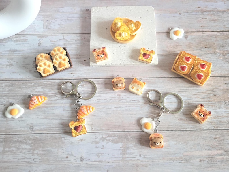 Puede incluir: Una variedad de colgantes y llaveros en miniatura con tem&aacute;tica de comida. Incluye tostadas con mermelada en forma de coraz&oacute;n, tostadas con forma de oso, cruasanes y colgantes con forma de huevo. Algunos colgantes est&aacute;n unidos a llaveros plateados.