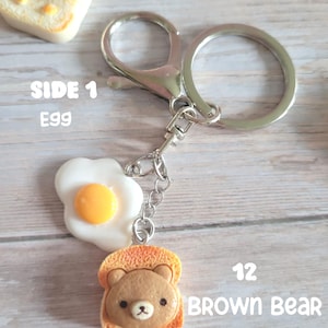 Puede incluir: Un llavero plateado con un huevo frito y una rebanada de pan tostado con la cara de un oso pardo. El texto "12 BROWN BEAR" es visible. Un dije separado muestra una rebanada de pan tostado con la cara de un oso sonriente. El texto "SIDE 1 EGG" es visible.