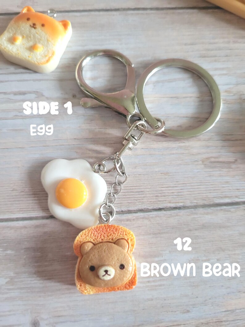 Puede incluir: Un llavero plateado con un huevo frito y una rebanada de pan tostado con la cara de un oso pardo. El texto "12 BROWN BEAR" es visible. Un dije separado muestra una rebanada de pan tostado con la cara de un oso sonriente. El texto "SIDE 1 EGG" es visible.
